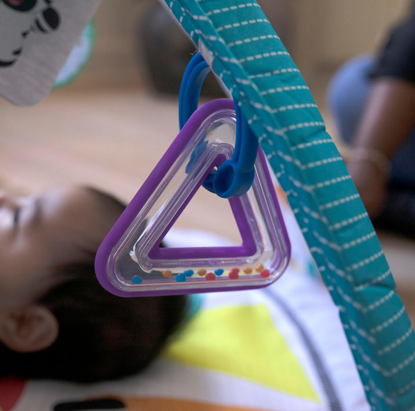 Gros plan du hochet triangle violet avec billes colorées suspendu à l'arche rayée turquoise du tapis d'éveil au-dessus du bébé