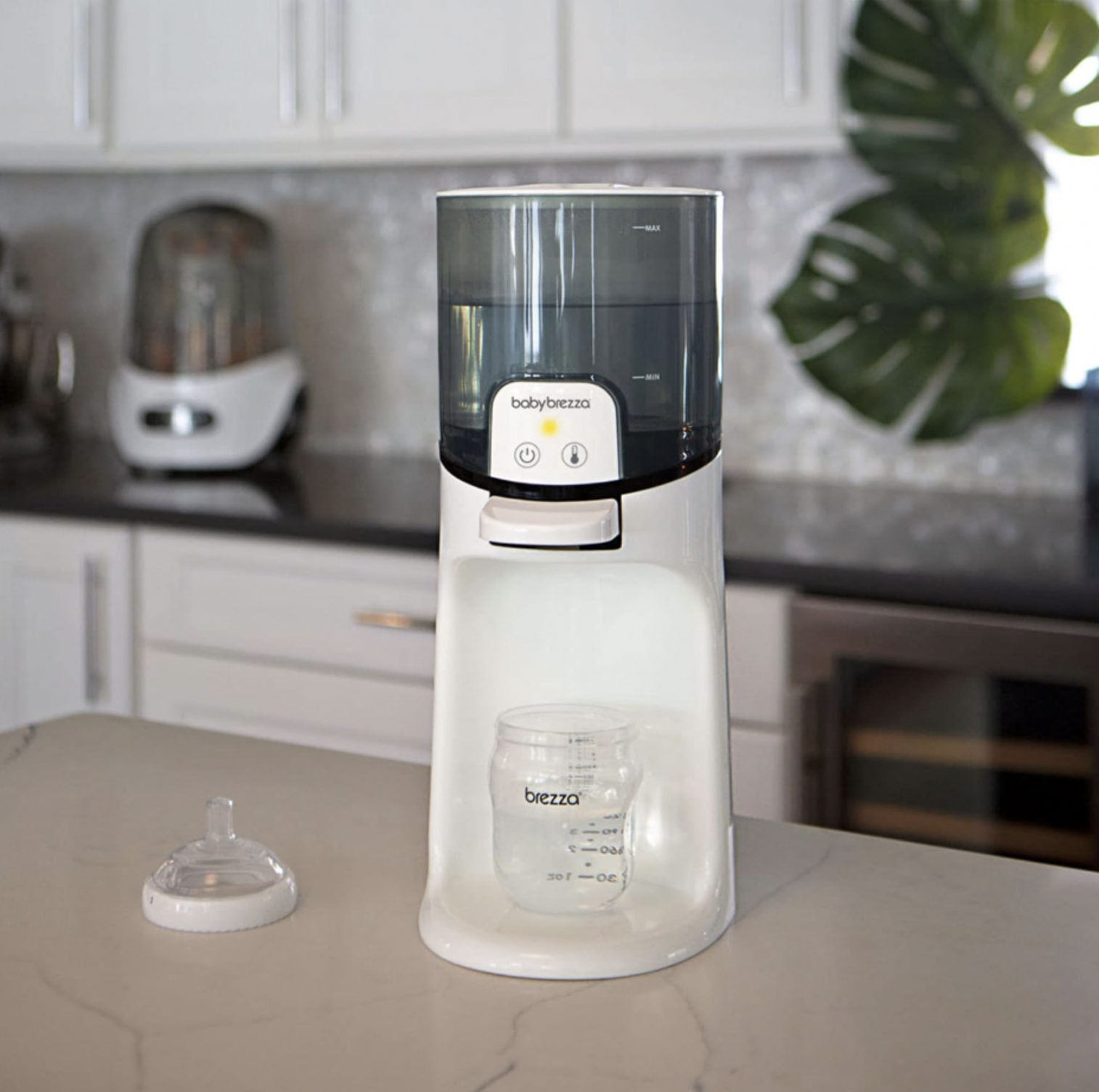 Chauffe-biberon instantané Baby Brezza sur un comptoir de cuisine en quartz blanc à côté d'une tétine de biberon avec feuille de monstera et armoires