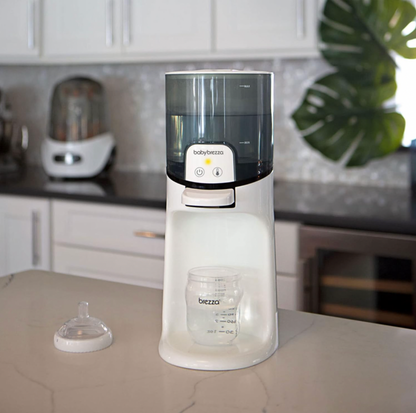 Chauffe-biberon instantané Baby Brezza sur un comptoir de cuisine en quartz blanc à côté d'une tétine de biberon avec feuille de monstera et armoires