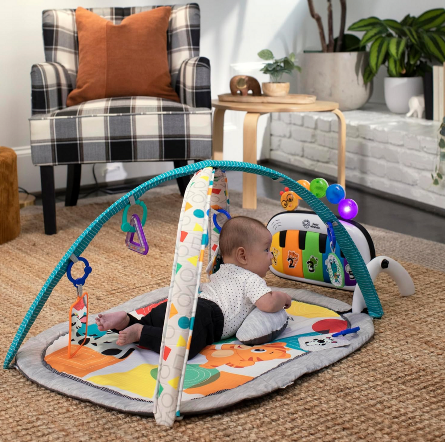 Bébé assis jouant sur le tapis d'éveil Baby Einstein avec arche turquoise et panneau piano dans un salon avec fauteuil à carreaux et plantes