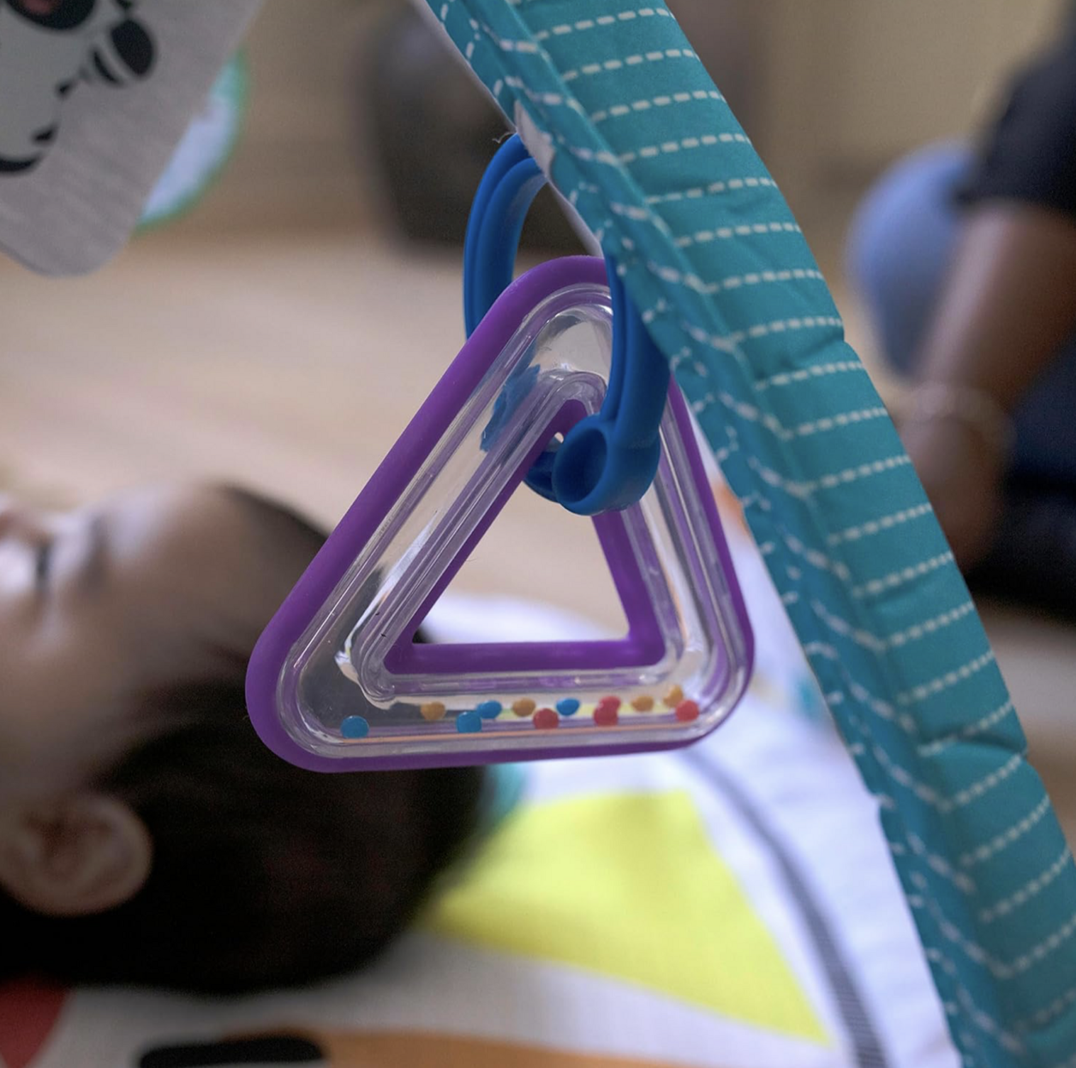 Gros plan du hochet triangle violet avec billes colorées suspendu à l'arche rayée turquoise du tapis d'éveil au-dessus du bébé