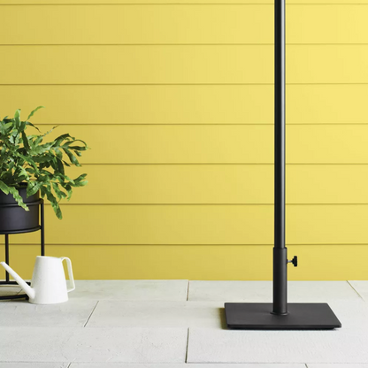 Base carrée noire de parasol sur un patio avec mur en lattes jaunes, arrosoir et plante en pot