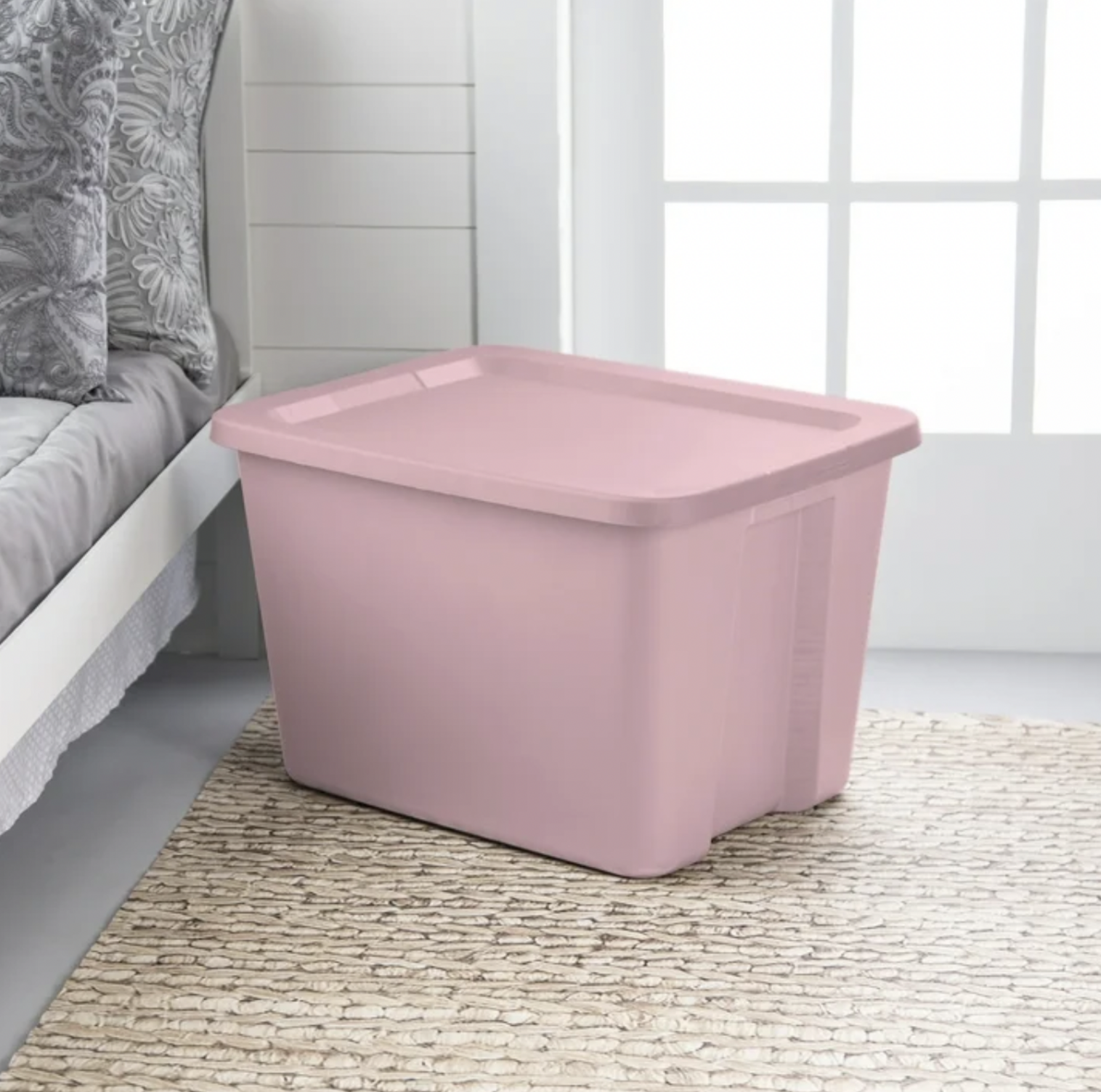 Boîte de rangement rose pâle posée sur un tapis tressé près d'un lit dans une chambre ensoleillée