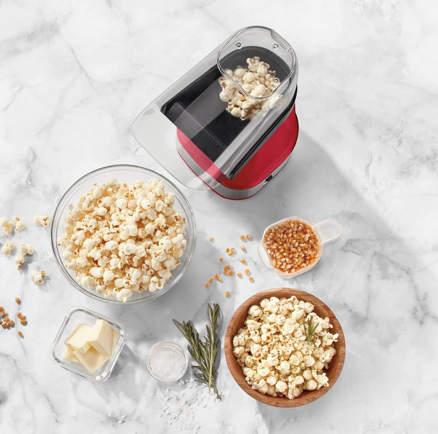 Vue de dessus de la machine à popcorn Cuisinart rouge avec maïs soufflé, grains, beurre et romarin sur comptoir en marbre
