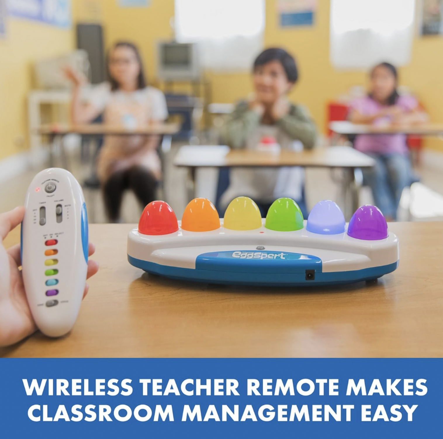 Main tenant la télécommande sans fil de l'enseignant devant la base de buzzer arc-en-ciel Eggspert avec élèves aux bureaux en arrière-plan, mention Wireless Teacher Remote
