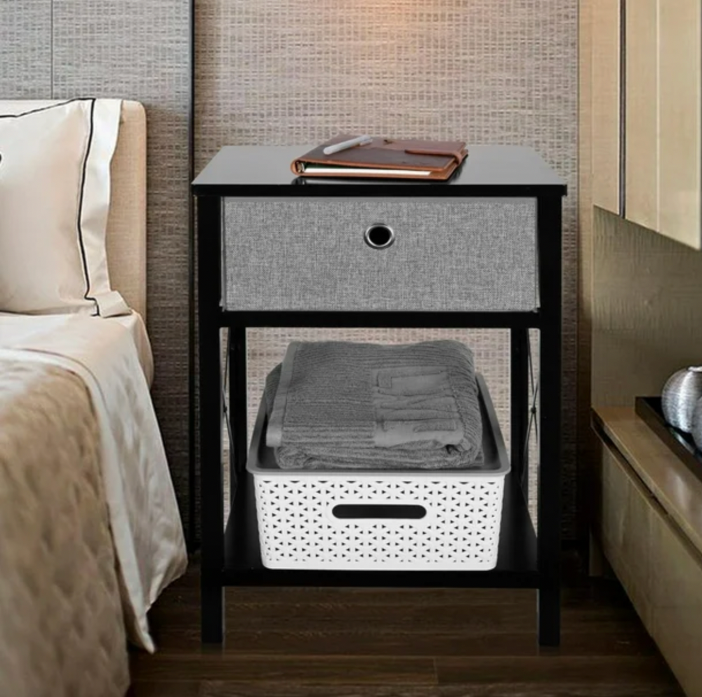 Table d'appoint noire à côté d'un lit avec livre en cuir sur le dessus, tiroir gris et serviettes pliées dans un panier blanc sur la tablette inférieure