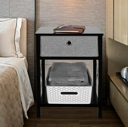 Table d'appoint noire à côté d'un lit avec livre en cuir sur le dessus, tiroir gris et serviettes pliées dans un panier blanc sur la tablette inférieure