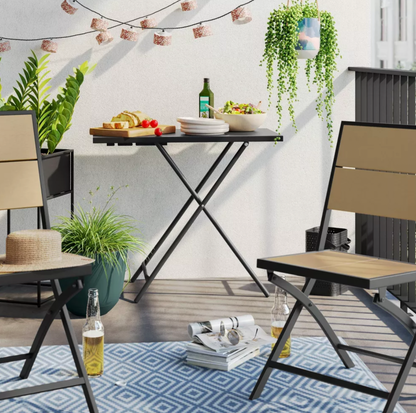 Table d'appoint pliante noire mise en scène sur un patio avec chaises à lattes de bois, plantes, guirlandes et planche à pain