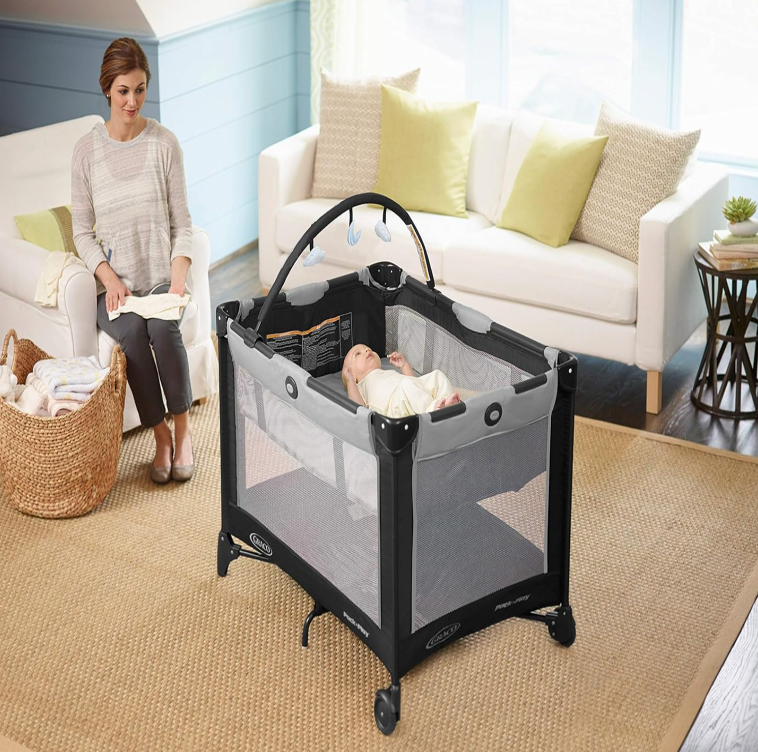 Bébé reposant dans la bassinette du parc Graco gris dans un salon avec canapé blanc et maman assise à côté