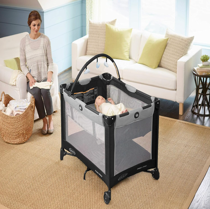 Bébé reposant dans la bassinette du parc Graco gris dans un salon avec canapé blanc et maman assise à côté