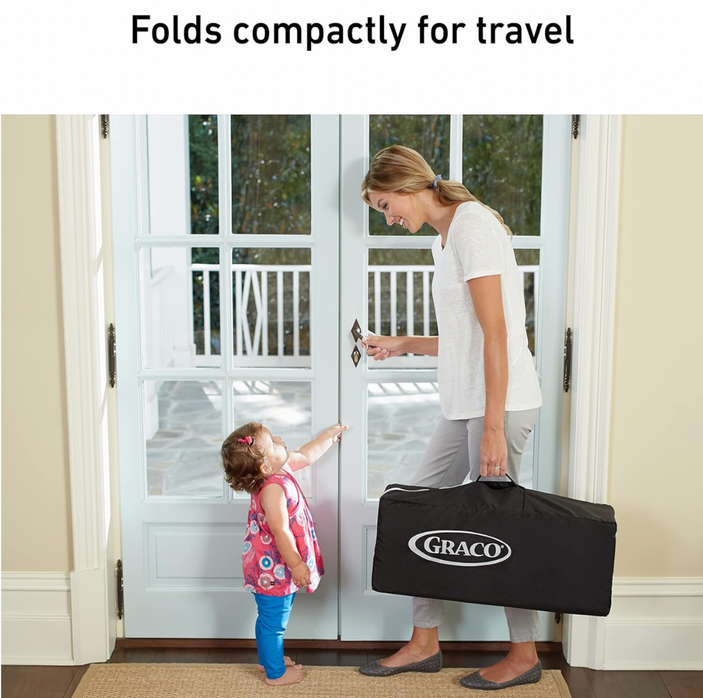 Maman tenant le parc Graco replié dans son sac de transport noir avec bambin à la porte d'entrée, mention Se replie de façon compacte pour le voyage