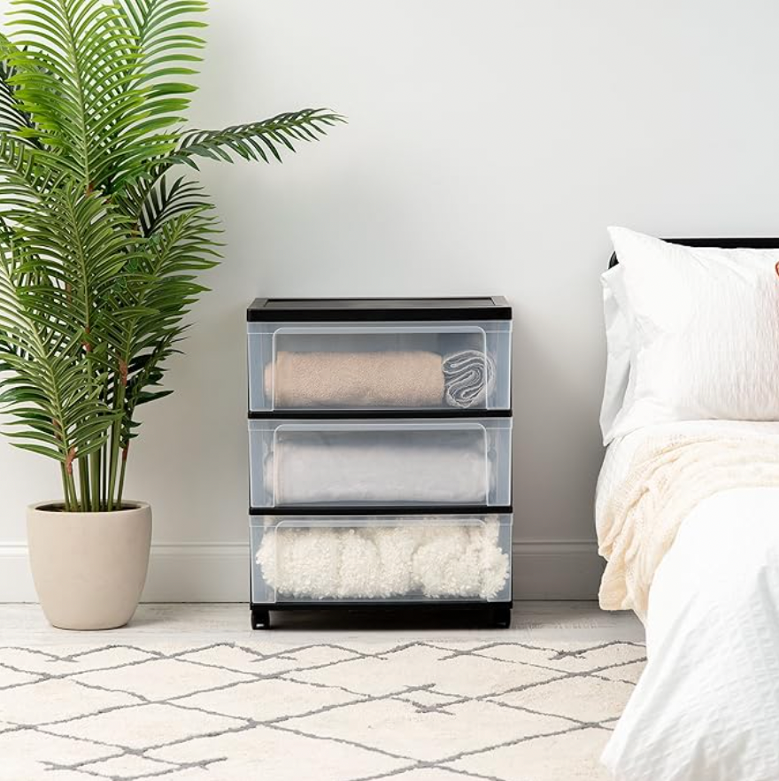 Chariot de rangement en plastique gris à 3 tiroirs dans une chambre avec serviettes pliées à côté d'un lit et d'une plante à feuillage vert