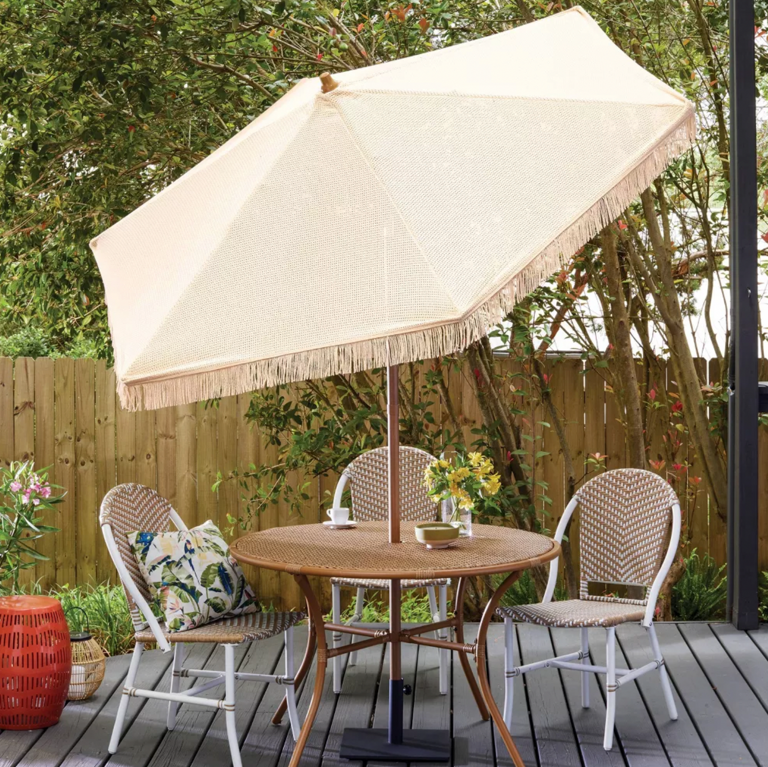 Parasol en macramé beige à franges ombrageant un ensemble repas de patio en rotin sur terrasse en bois avec coussins et fleurs