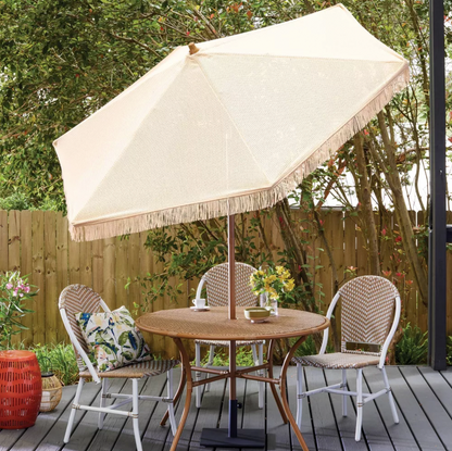 Parasol en macramé beige à franges ombrageant un ensemble repas de patio en rotin sur terrasse en bois avec coussins et fleurs