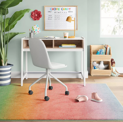 Coin bureau d'enfant avec bureau blanc, chaise, tableau des tâches et tapis touffeté en dégradé arc-en-ciel couvrant le sol avec chaussures et plante