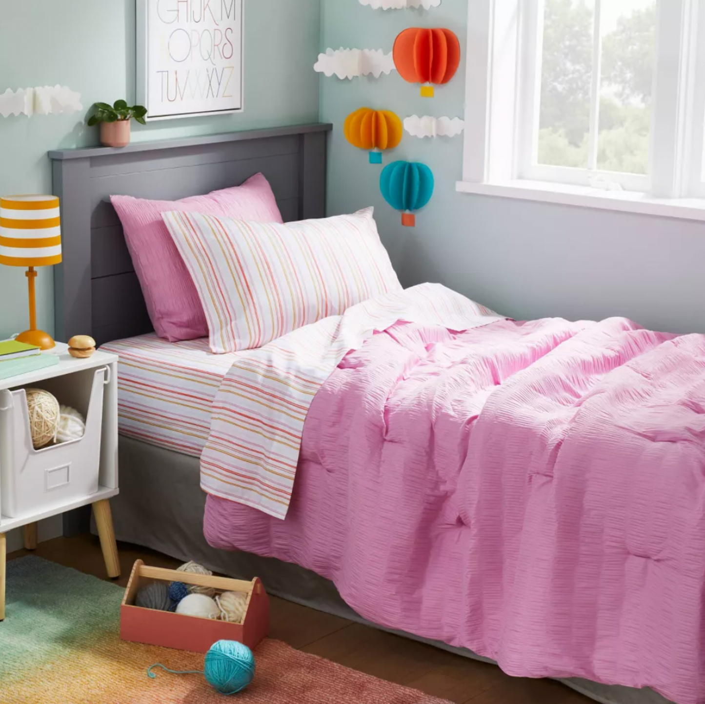 Coin de chambre d'enfant avec literie matelassée rose, coussin à rayures et tapis en dégradé arc-en-ciel visible au sol près d'une table de chevet blanche