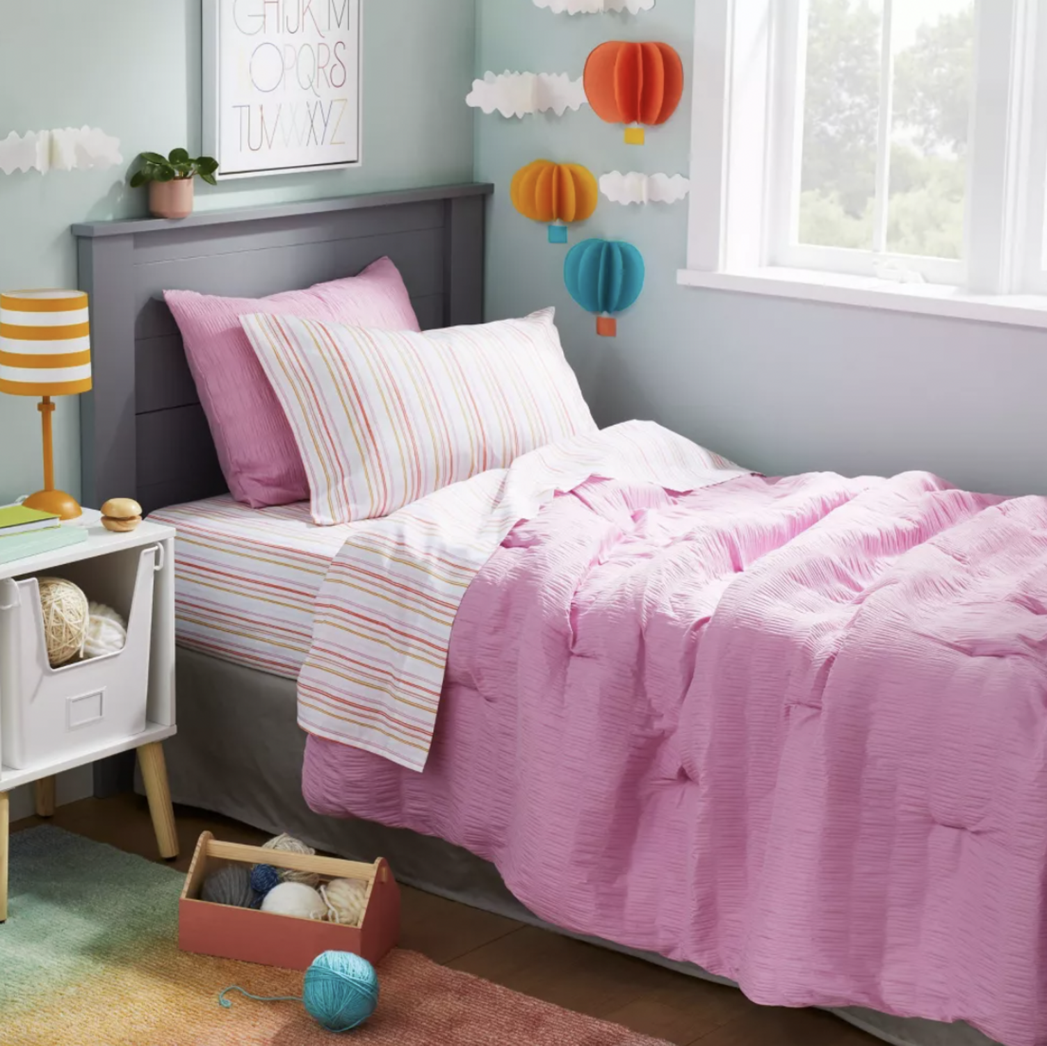 Coin de chambre d'enfant avec literie matelassée rose, coussin à rayures et tapis en dégradé arc-en-ciel visible au sol près d'une table de chevet blanche
