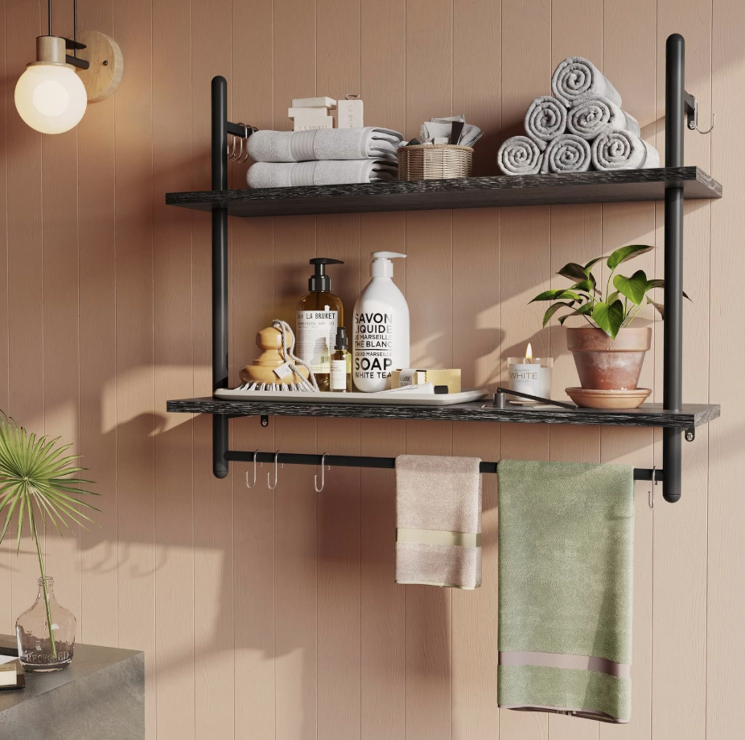 Étagère à tuyaux noire à deux étages dans une salle de bain avec serviettes roulées, distributeurs de savon, bougie et serviette suspendue à la barre