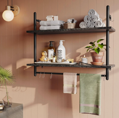 Étagère à tuyaux noire à deux étages dans une salle de bain avec serviettes roulées, distributeurs de savon, bougie et serviette suspendue à la barre