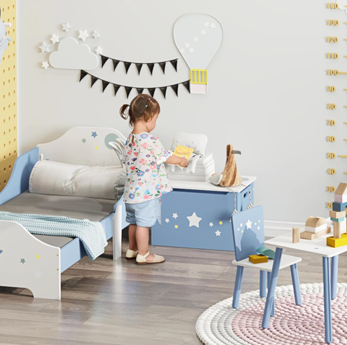 Fillette plaçant un livre sur le coffre à jouets bleu dans une chambre d'enfant avec décor mural de nuage et ballon et tableau perforé