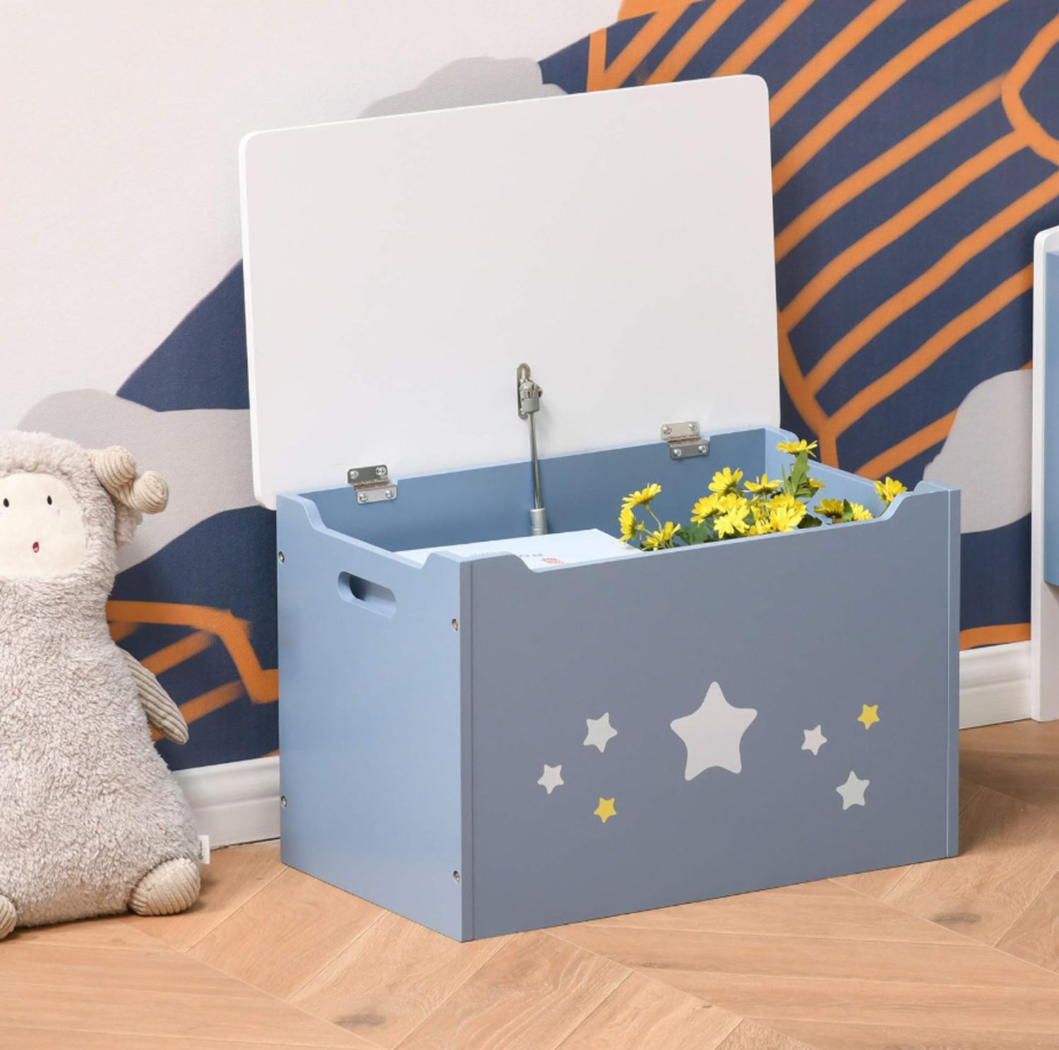 Coffre à jouets Qaba bleu avec couvercle blanc ouvert révélant des marguerites jaunes à l'intérieur, à côté d'une peluche agneau dans une salle de jeux avec mur à motif de nuages
