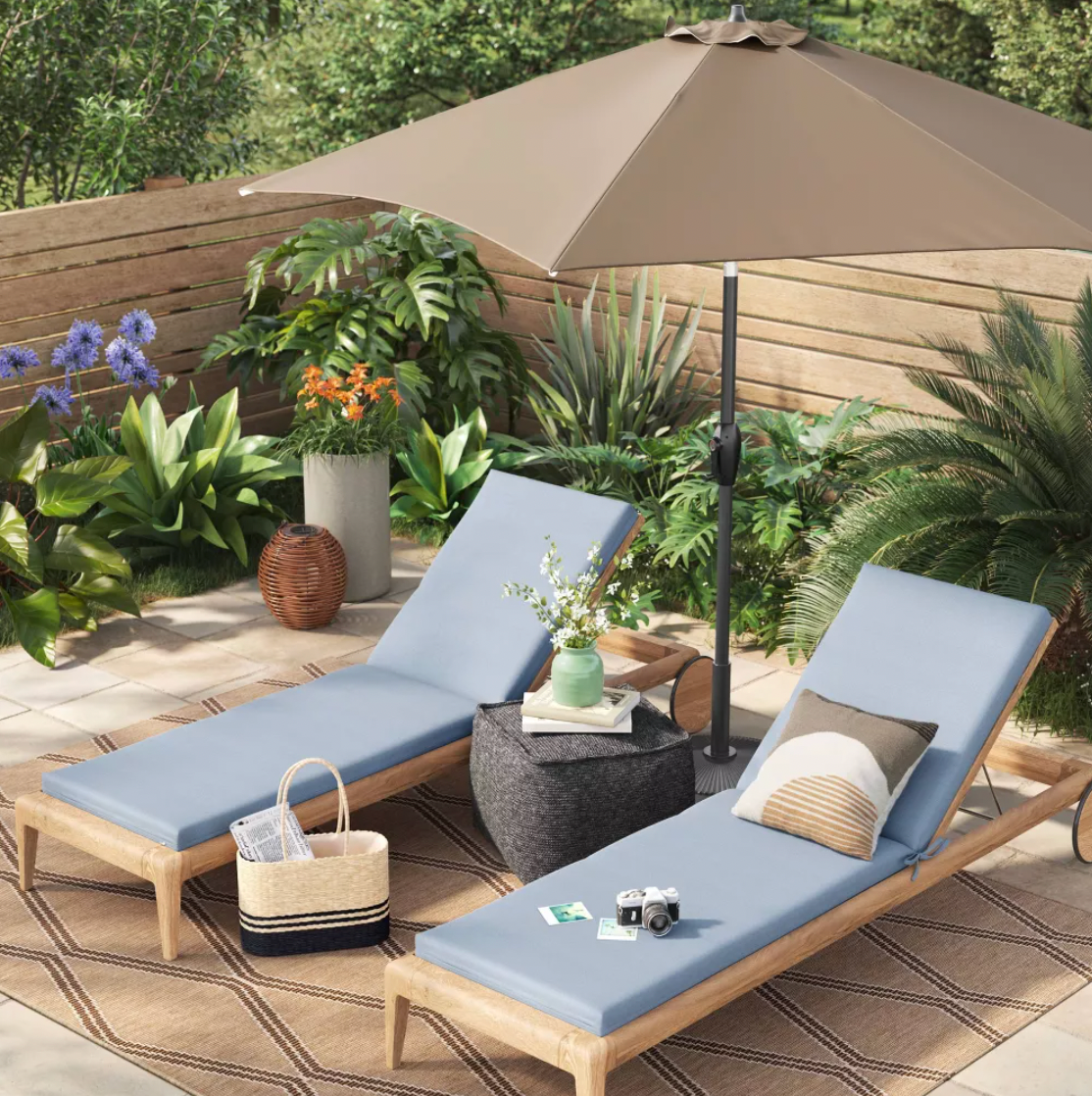 Brown patio umbrella shading two wood chaise lounges with blue cushions and side table on outdoor terrace