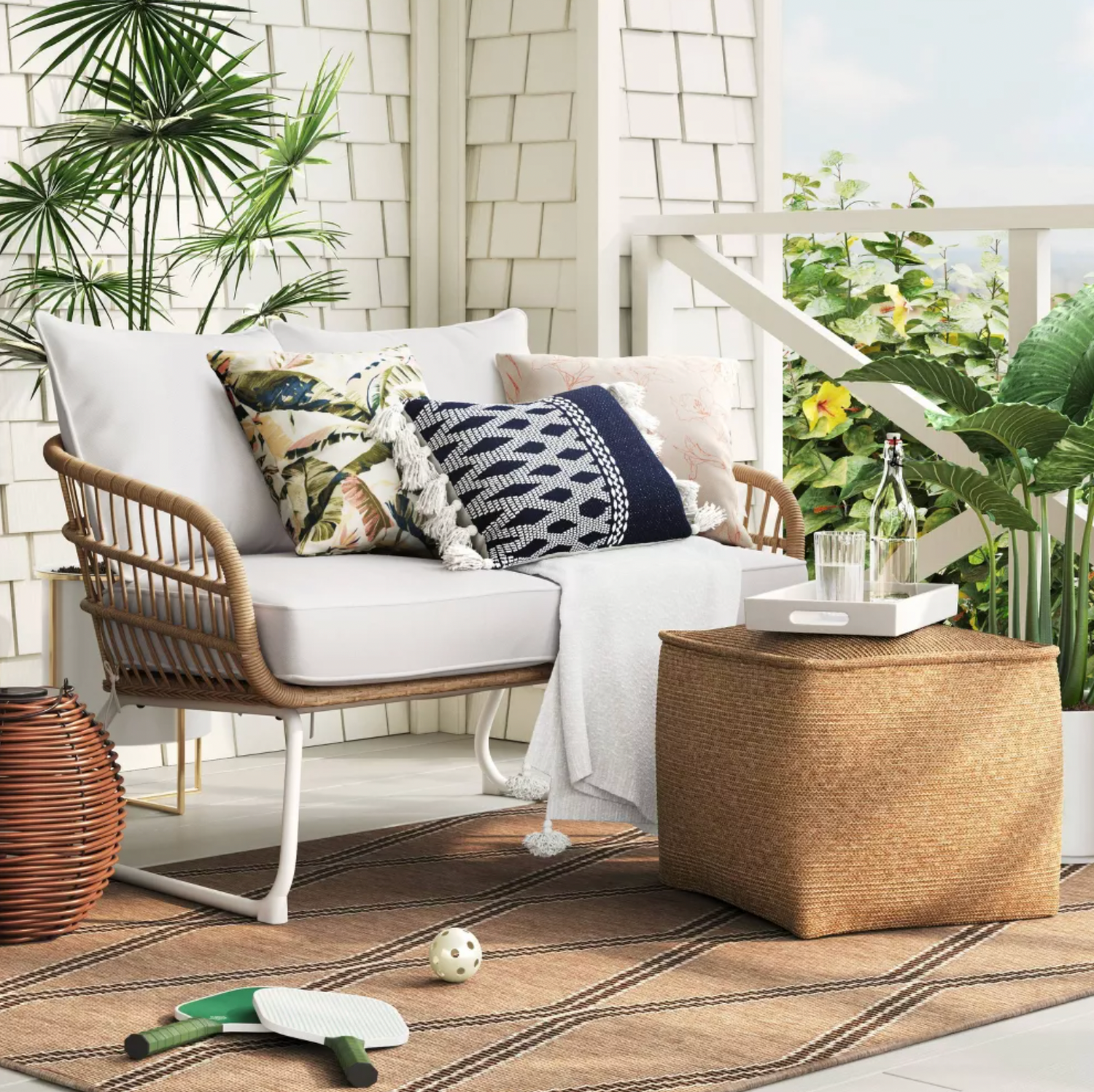 Pouf tricoté tan utilisé comme table d'appoint sur une véranda couverte avec causeuse en rotin, coussins et palmiers