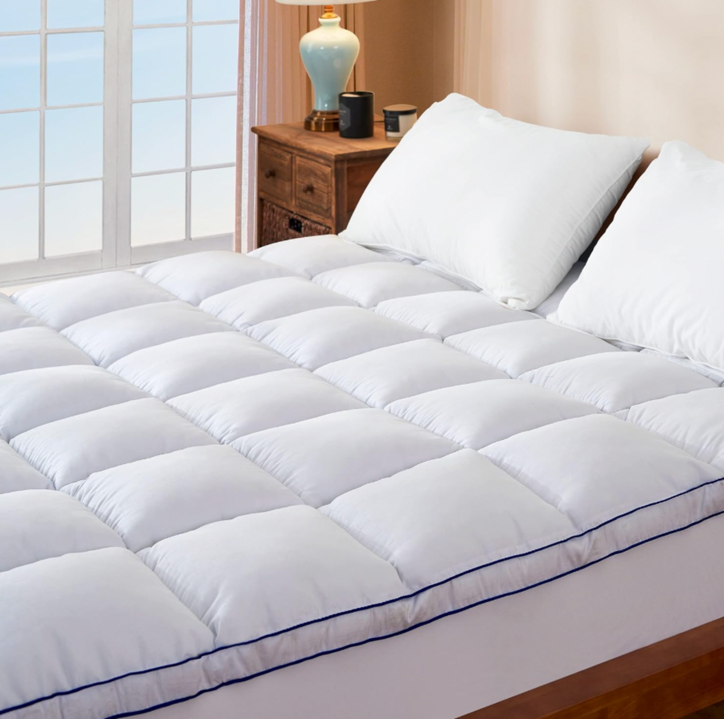 Surmatelas matelassé blanc installé sur un lit avec oreillers blancs dans une chambre avec table de chevet en bois et lampe de table