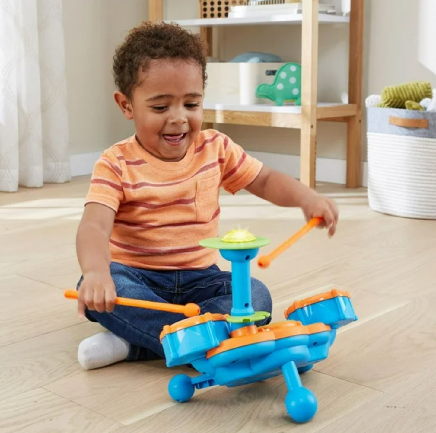 Boy in striped shirt playing VTech Kidibeats blue drum set with orange drumsticks on living room floor