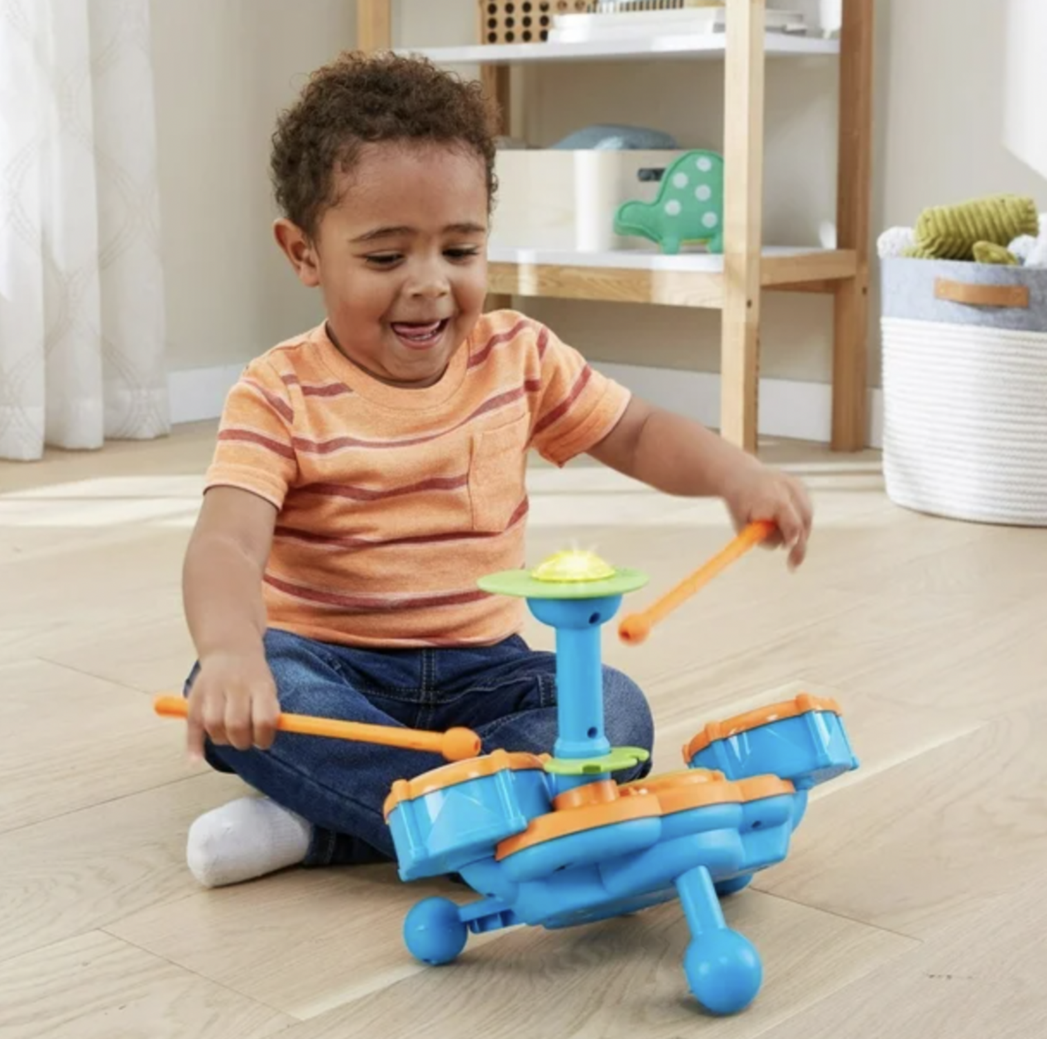 Boy in striped shirt playing VTech Kidibeats blue drum set with orange drumsticks on living room floor