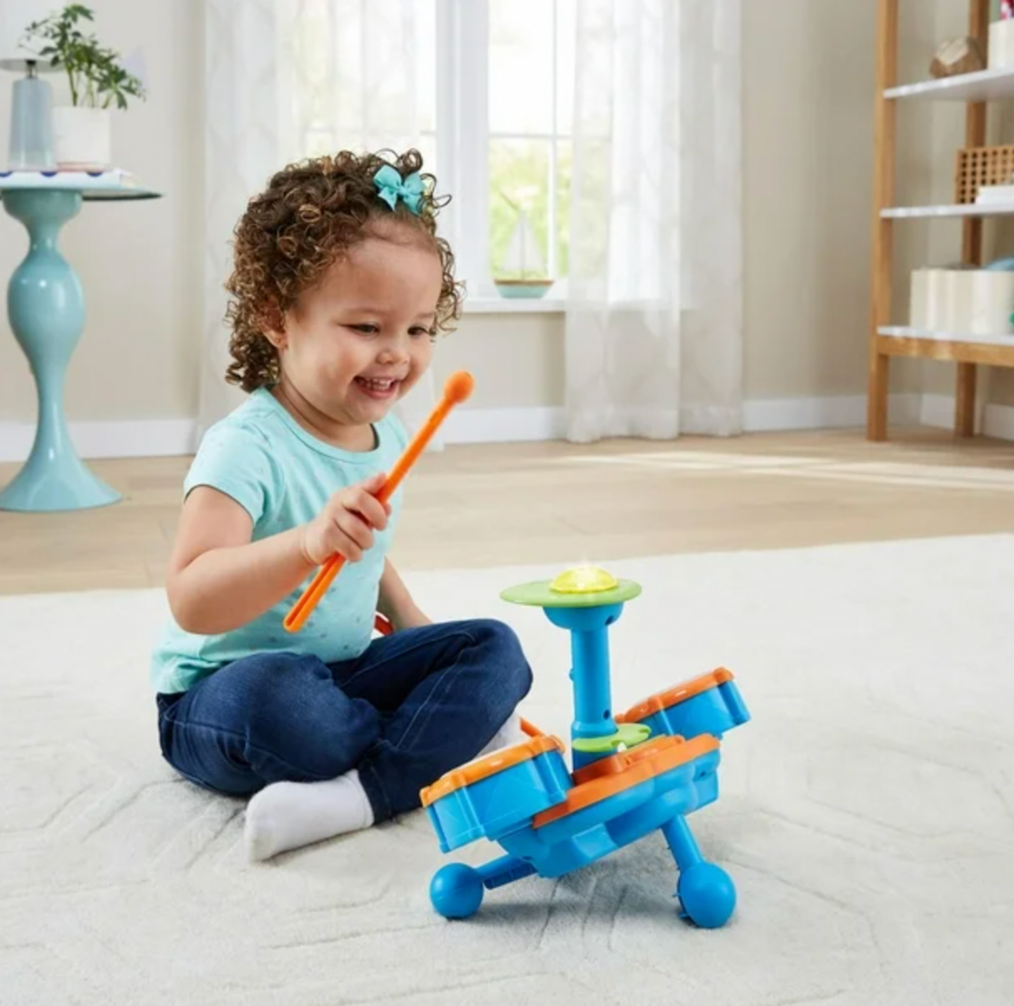 Toddler girl with bow playing VTech Kidibeats drum set with orange drumstick in bright living room