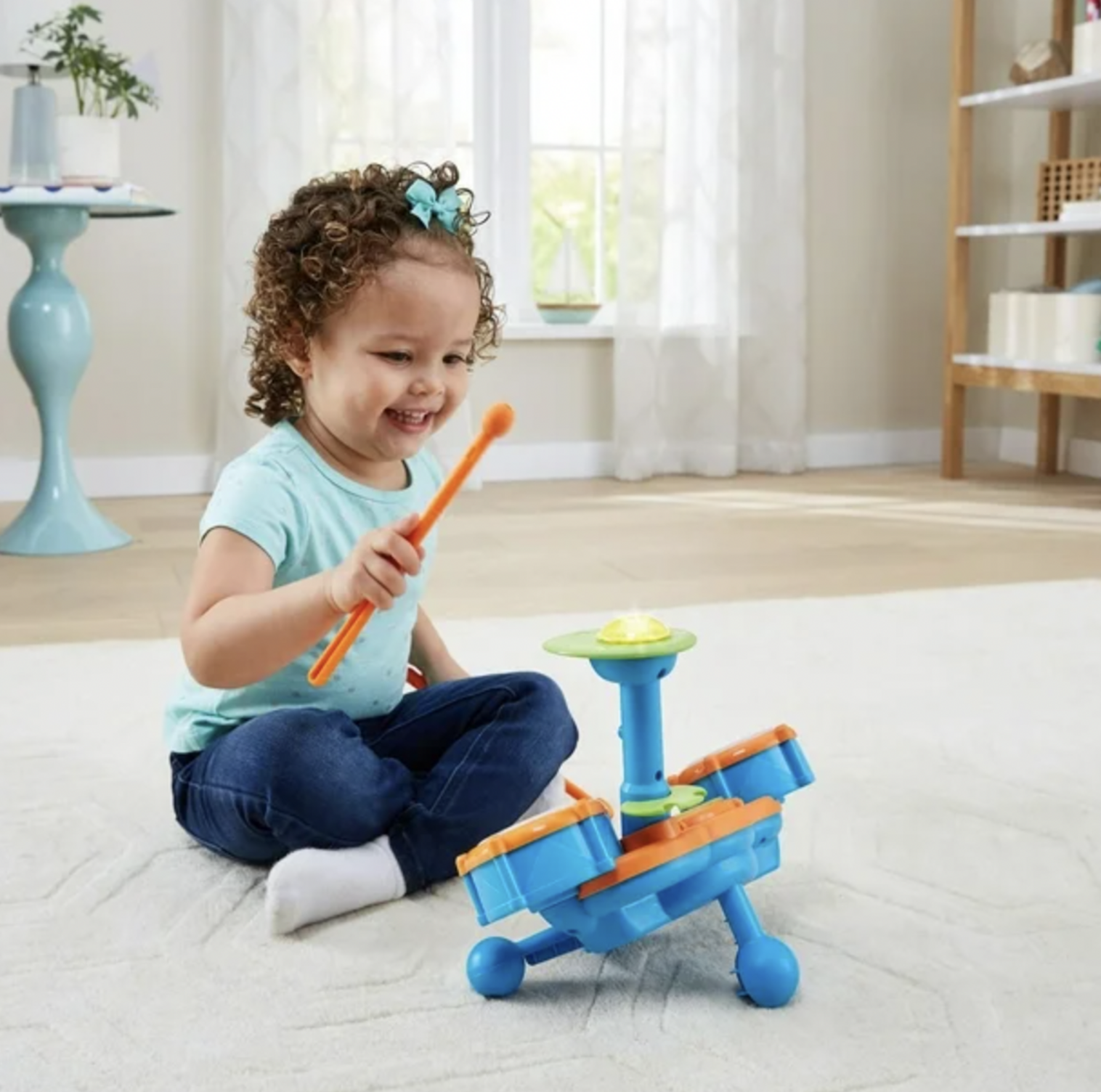 Toddler girl with bow playing VTech Kidibeats drum set with orange drumstick in bright living room